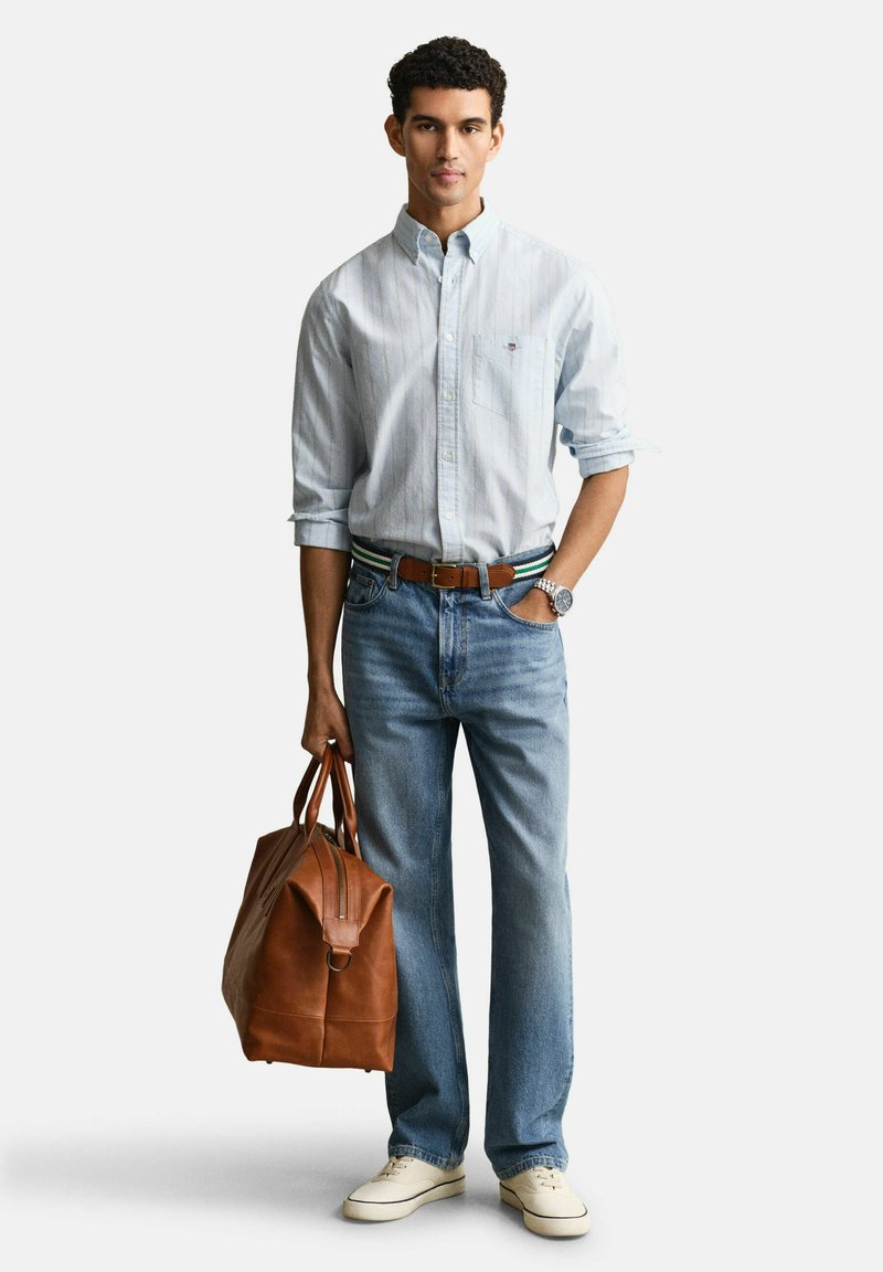 Uomo che indossa una camicia azzurra a righe con bottoni, jeans blu, sneakers bianche, cintura marrone, che tiene una grande borsa in pelle marrone.