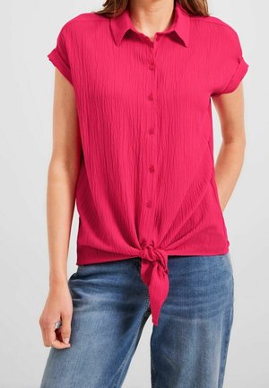 Blouse rose à manches courtes avec un tissu texturé et un détail de nœud à l'avant. Dotée d'un col boutonné et de manches retroussées. Associée à un jean bleu.