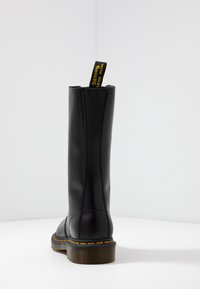Bota de cuero negro hasta la rodilla con textura suave, acento amarillo en el tirador y costura visible en la costura trasera. Suela de goma.