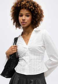 Γυναίκα με σγουρά μαλλιά, φορώντας εφαρμοστό λευκό πουκάμισο με κουμπιά και παντελόνι με λεπτές ρίγες, κρατώντας μια μαύρη τσάντα ώμου με υφή.