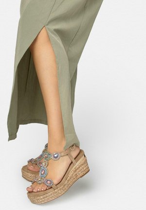 Piernas con sandalias de cuña beige con adornos de piedras preciosas multicolores, vestidas con una falda oliva clara con apertura, sobre un fondo liso.