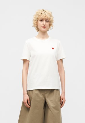 Jeune personne aux cheveux blonds bouclés, portant un t-shirt blanc avec un petit cœur rouge sur la poitrine et un pantalon beige ample.
