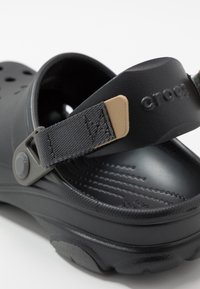 Sabot Crocs noir avec bride arrière en tissu gris réglable et accent beige, semelle texturée et trous d'aération au niveau des orteils.