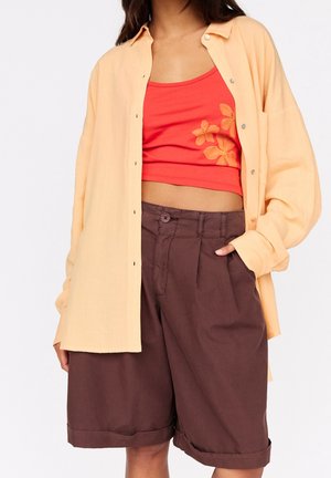 Femme portant une chemise ample couleur pêche boutonnée sur un haut court rouge à motifs floraux, associée à un short marron taille haute mi-longueur, une main dans la poche.