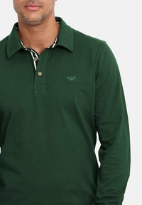 Camisa polo de manga larga en verde oscuro, fabricada en algodón, con un cuello clásico, tres botones y un sutil logo bordado en el pecho.
