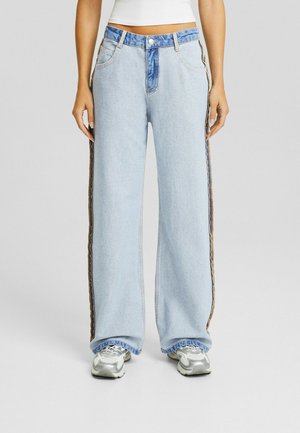 Jeans en denim bleu clair à jambes larges, avec une ceinture contrastante bleue et des bandes latérales texturées marron. Coupe décontractée avec un ourlet brut.