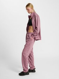 Femme blonde en survêtement mauve avec un soutien-gorge de sport noir et des baskets noires, debout, les mains dans les poches, sur un fond uni.