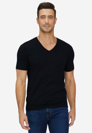 Mann mit dunklen Haaren, der ein schwarzes kurzärmeliges V-Ausschnitt-Shirt und blaue Jeans trägt, steht vor weißem Hintergrund.