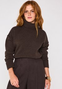 Maglione a collo alto marrone cioccolato con texture a coste e spalle scese, abbinato a pantaloni scuri coordinati e bracciali dorati.