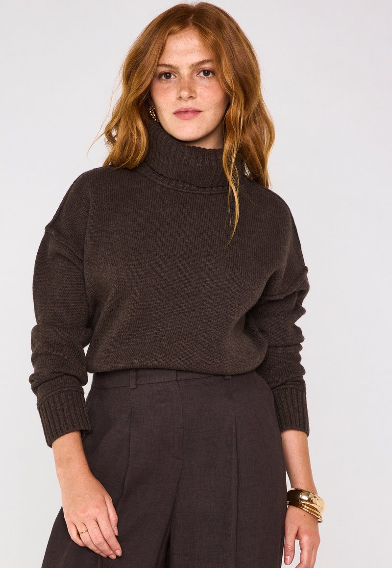 Maglione a collo alto marrone cioccolato con texture a coste e spalle scese, abbinato a pantaloni scuri coordinati e bracciali dorati.