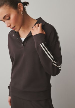 Femeie atingând gulerul unui top întunecat cu mâneci lungi, cu dungi albe pe mâneci, purtând cercei cu cerc auriu și ear cuff, privind în jos.