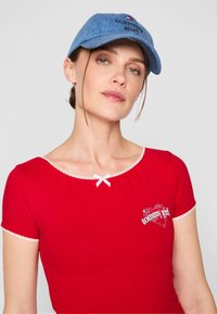 Rød topp med korte ermer, blondekant og en hvit sløyfe ved halsen, med en hjerteformet "tommy girl" grafikk i hvitt og rødt.