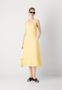 Rochie fără mâneci din in galben, cu decolteu în V, lungime până la genunchi, buzunare laterale și un detaliu subtil de design. Asortată cu sandale negre tip slide.