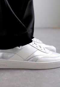 Baskets en cuir blanc avec une bande accent texturée en argent, dotées d'un design à lacets et de semelles en caoutchouc. Le matériau de base est lisse avec des détails de couture.