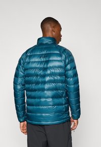 Fyldig teal pufferjakke med vandret syninger, høj krave og et lille logo på ryggen. Glat, skinnende tekstur og isoleret design.