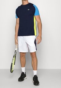 Námořnicky modré sportovní tričko s modrými a žlutými bočními panely, doplněné bílými šortkami s námořnicky modrým akcentem. Model drží tenisovou raketu.