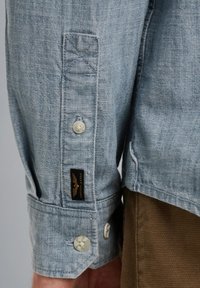 Camisa texturizada de color azul claro, con mangas largas, que presenta un puño abotonado y una etiqueta negra. Tejido suave y detalles de costura visibles.