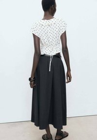 White blouse with black polka dots and ruffled collar, paired with a long black skirt. The outfit features a drawstring at the back and flat sandals.