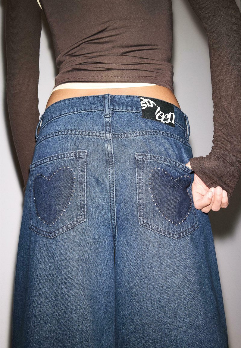 Personne portant un jean bleu taille haute avec des motifs de cœurs cloutés sur les poches arrière et une chemise marron à manches longues, main dans la poche.