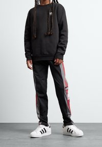 Person, der einen schwarzen Adidas-Pullover und schwarze Sporthosen mit roten und grauen Streifen trägt, steht auf einem grauen Boden vor einem weißen Hintergrund.