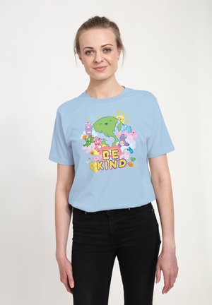 Femme portant une chemise bleu clair avec un dessin animé de la Terre, du soleil, des ours colorés, des fleurs, des cœurs, et le texte "SOYEZ GENTIL" à l'avant.