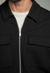 Sort lynlåstrøje med tekstureret vævning, to brystlommer og en sølvfarvet lynlås. Båret over en lysegrå crewneck-trøje.