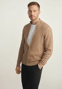 Sweter z dzianiny w jasnym beżu z wzorem w warkocze, zapinany na zamek błyskawiczny, z wysokim kołnierzem. Nosi się go na białej koszuli, w połączeniu z czarnymi spodniami.