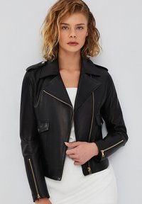 Zwarte leren motorjack met grote revers, asymmetrische rits en zakken. Heeft gouden accenten en een getailleerd ontwerp.