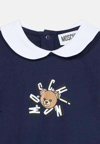 Temno modra obleka z belim ovratnikom v stilu Peter Pan, centralna grafika medvedka obkrožena z raztresenimi belimi črkami, ki tvorijo napis MOSCHINO.