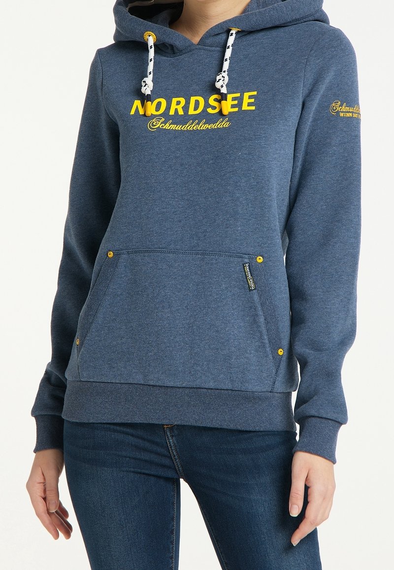 Žena oblečená v modré mikině s žlutým nápisem "NORDSEE" a bílými tkaničkami, kombinovaná s úzkými tmavě modrými džíny, stojící na bílém pozadí.