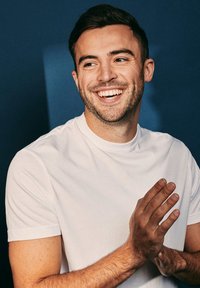 Wit T-shirt met korte mouwen; katoenen materiaal; model met kort donker haar; handen samengevouwen, glimlachend tegen een donkerblauwe achtergrond.