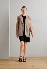 Beige bleiseri mustan mekon päällä, yhdistettynä mustiin korkokenkiin. Sileä kangas, rakenteellinen muotoilu, yksinkertainen nappidetail, ja suuret taskut.
