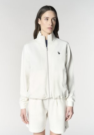 Donna che indossa giacca bianca con zip e pantaloncini abbinati con piccolo logo nero a forma di cavallo, in piedi su sfondo chiaro uniforme.