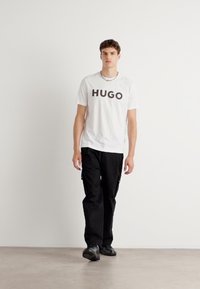 Fehér póló, rajta vastag fekete betűkkel a "HUGO" felirattal, fekete cargos nadrággal és fekete cipővel, egy semleges padlón áll.