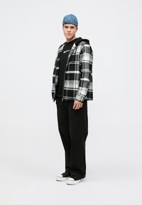 Chaqueta de franela a cuadros gris y negra sobre una camiseta negra, combinada con pantalones negros y zapatillas negras. El modelo lleva una gorra azul claro.