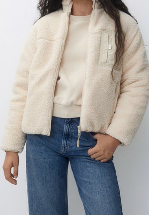 Kvinde iført cremefarvet fleecejakke med lynlås, beige skjorte og blå jeans, stående med den ene hånd i lommen.