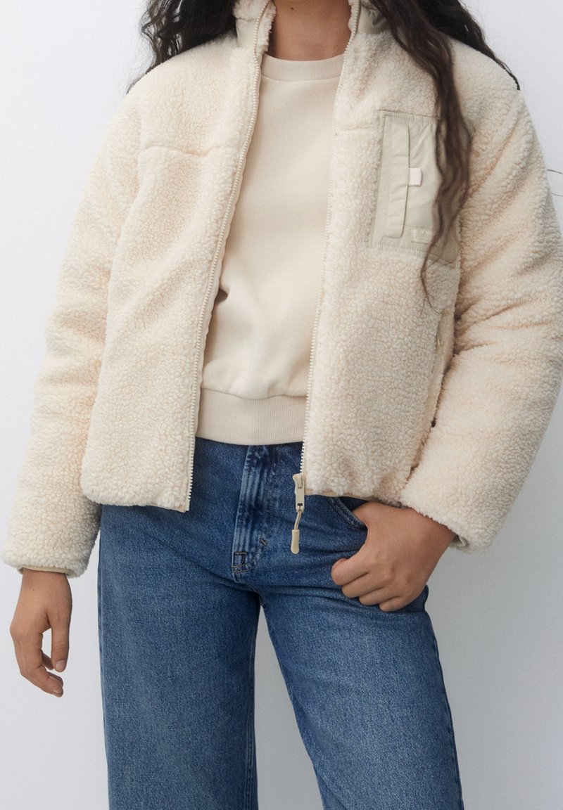 Femme portant une veste polaire crème avec fermeture éclair, chemise beige et jean bleu, debout, une main dans la poche.