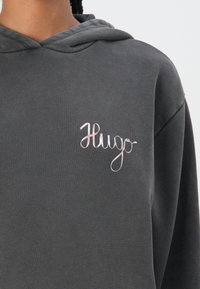 Haut à capuche gris en matériau doux, avec un cordon de serrage à la capuche et un logo "Hugo" brodé en rose sur la poitrine.