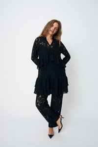 Outfit in pizzo nero con una tunica a strati con volant e pantaloni coordinati. Il tessuto presenta intricati motivi floreali e una trama semi-trasparente.