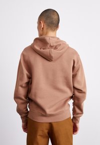 Sudadera marrón con capucha con cordón, textura suave, puños y dobladillo acanalados. Llevada con pantalones beige, vista desde atrás. Diseño simple y limpio.