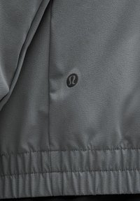 Veste de sport grise avec un tissu texturé, un ourlet élastique et un petit logo noir. Surface lisse avec un minimum de détails.