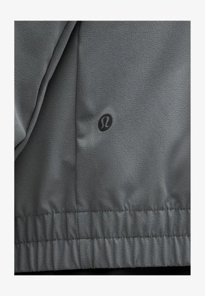 Chaqueta atlética gris con un tejido texturizado, dobladillo elástico y un pequeño logotipo negro. Superficie suave con un detalle mínimo.