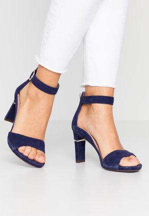 Marineblauwe suède hoge hakken met een open teen, enkelbandje en een rechthoekige hak, voorzien van een metallic accent nabij de hak.