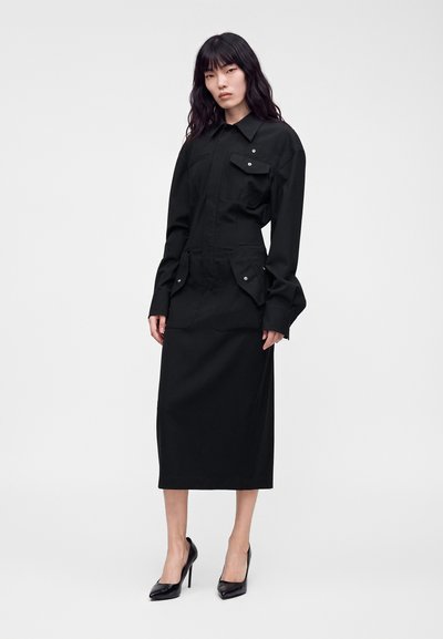 Femme aux longs cheveux noirs portant une robe-chemise noire à manches longues avec plusieurs poches et des chaussures noires à talons hauts, debout sur un fond blanc.