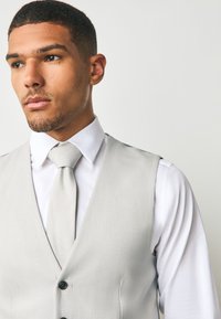 Gilet gris clair texturé avec un design en V prononcé, comportant une chemise habillée blanche et une cravate grise assortie. Boutons à la taille.