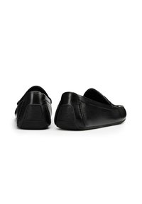 Zapatos negros de cuero sin cordones con un tacón bajo, puntera redondeada y suela texturizada. Presenta costuras decorativas a lo largo de la parte superior.