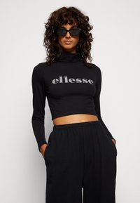 Juodas trumpas kaklo megztinis su pilku "ellesse" logotipu, derinamas su juodomis laisvomis kelnėmis. Tamsūs nepraleidžiantys saulės akiniai papildo įvaizdį.