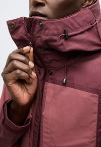 Bărbat cu piele închisă la culoare care închide fermoarul unei jachete bordo cu guler înalt și glugă reglabilă, arătând mâna și o parte din fața sa.