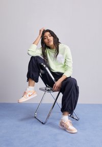 Sweat-shirt vert clair avec logo blanc, pantalon cargo marine, et baskets Nike orange pastel sur une chaise pliante chromée, sur un fond gris.