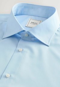 Hellblaues Hemd aus glattem Stoff, mit klassischem Kragen, weißen Knöpfen und einem Etikett, das "NEXT TAILORING" liest.